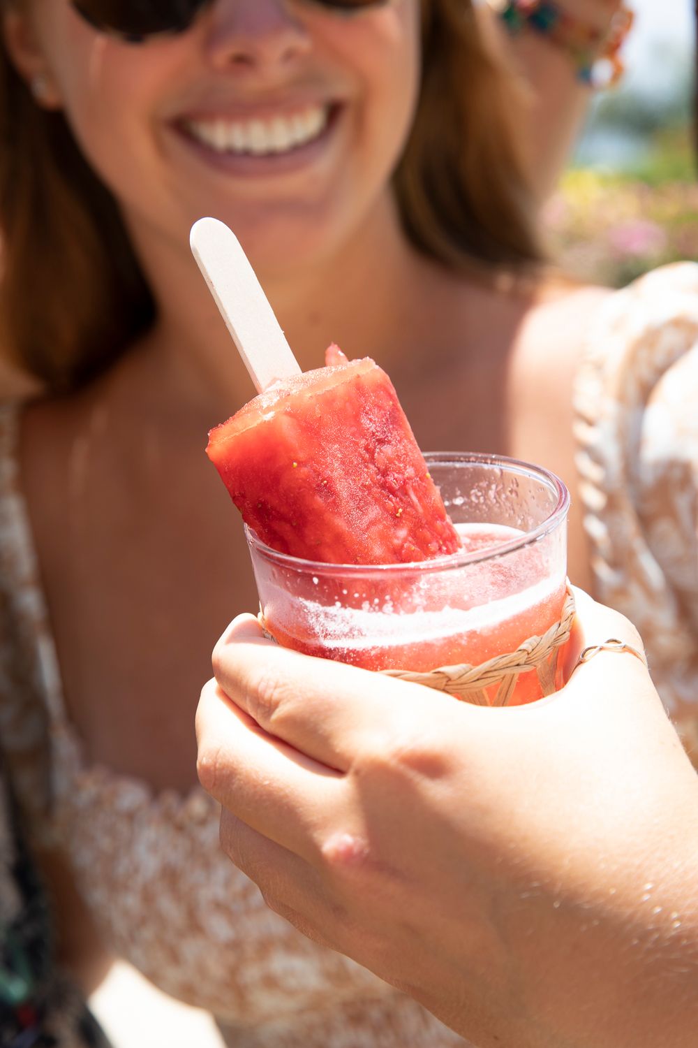 Woman smiling while holding a refreshing watermelon popsicle in a clear cup on a sunny day.