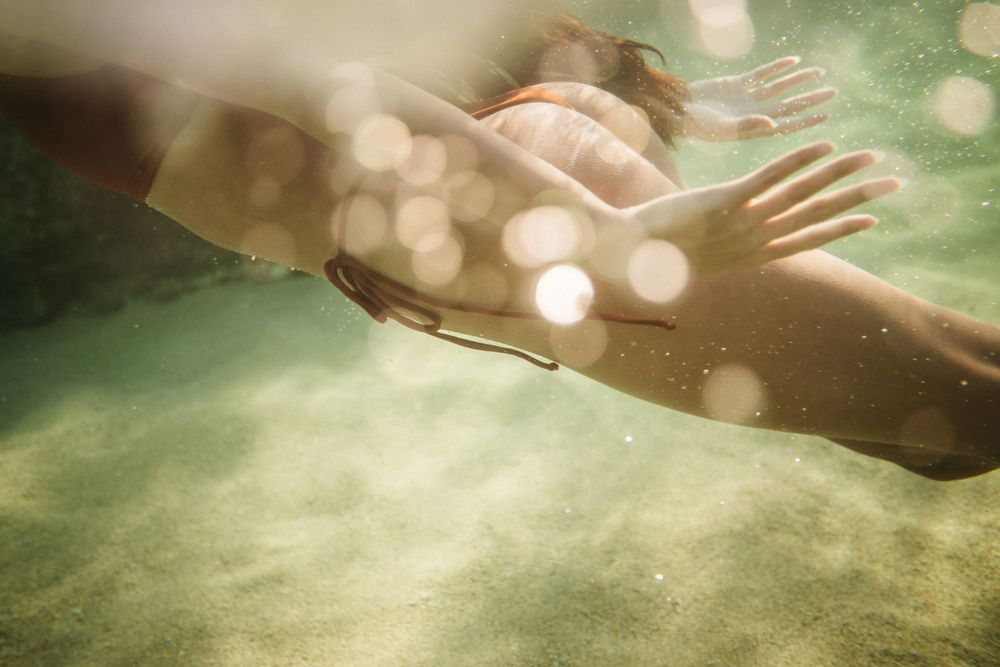 Person swimming underwater with sunlight filtering through the water, creating bokeh light effects on sandy bottom.
