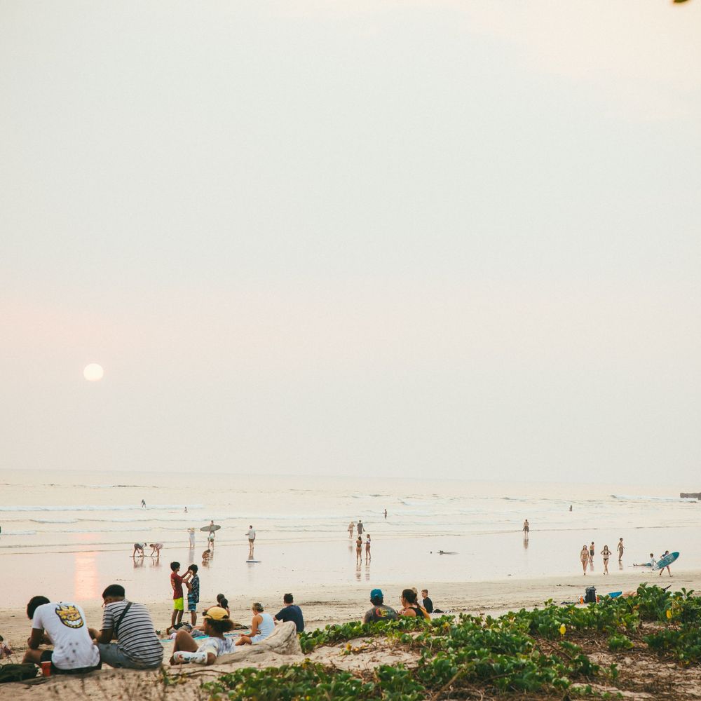 People relaxing on a sandy beach at sunset with calm ocean waves and a hazy golden sky in the background.