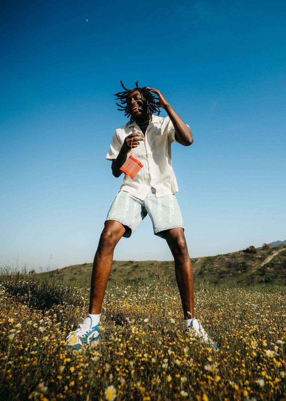 Young man with dreadlocks posing in a wildflower field, wearing white shorts and shirt, holding a red book under blue sky.