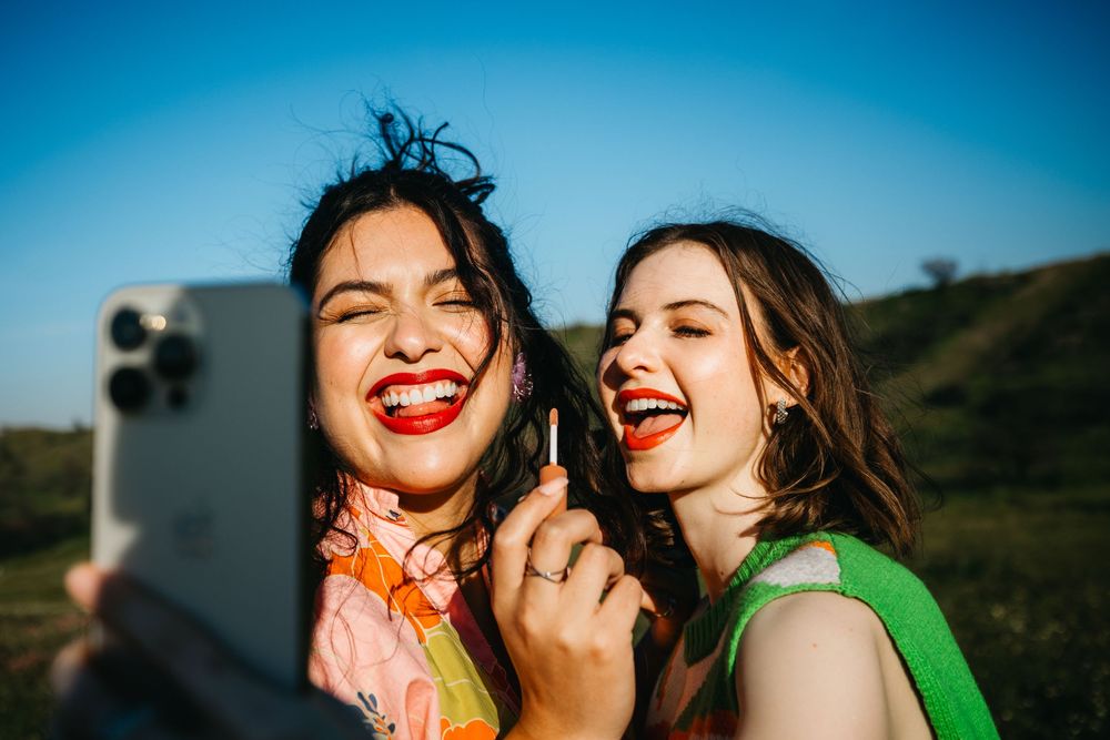 Two laughing women with red lips take a selfie outdoors on a sunny day, one holding a lipstick.