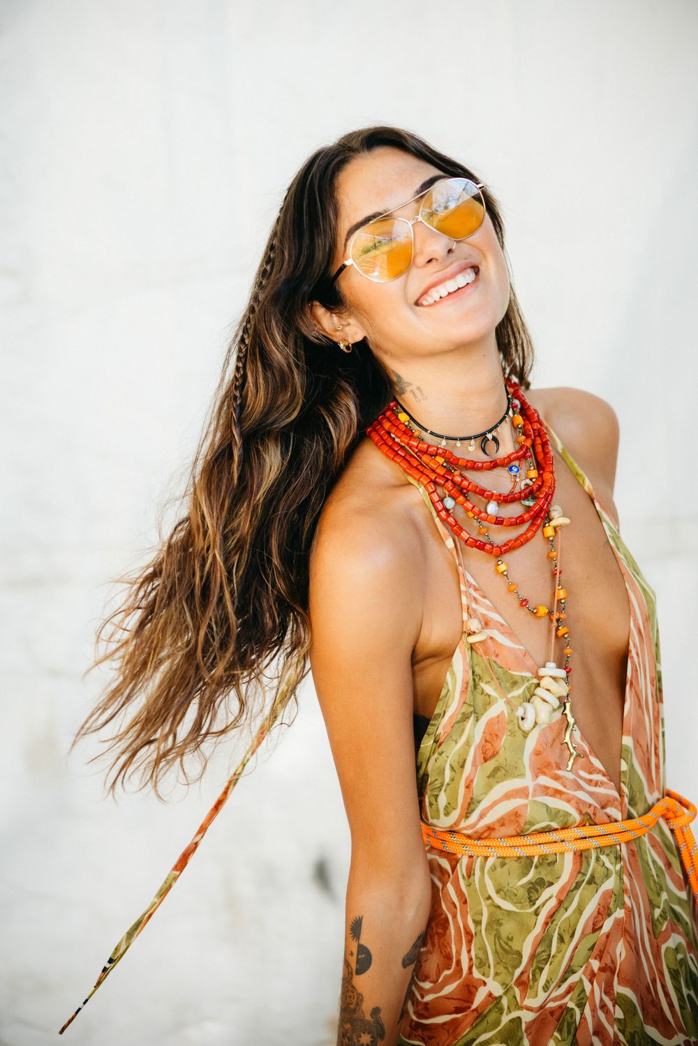 Smiling woman in boho style colorful dress, red beaded necklaces, yellow sunglasses, and long wavy hair outdoors.