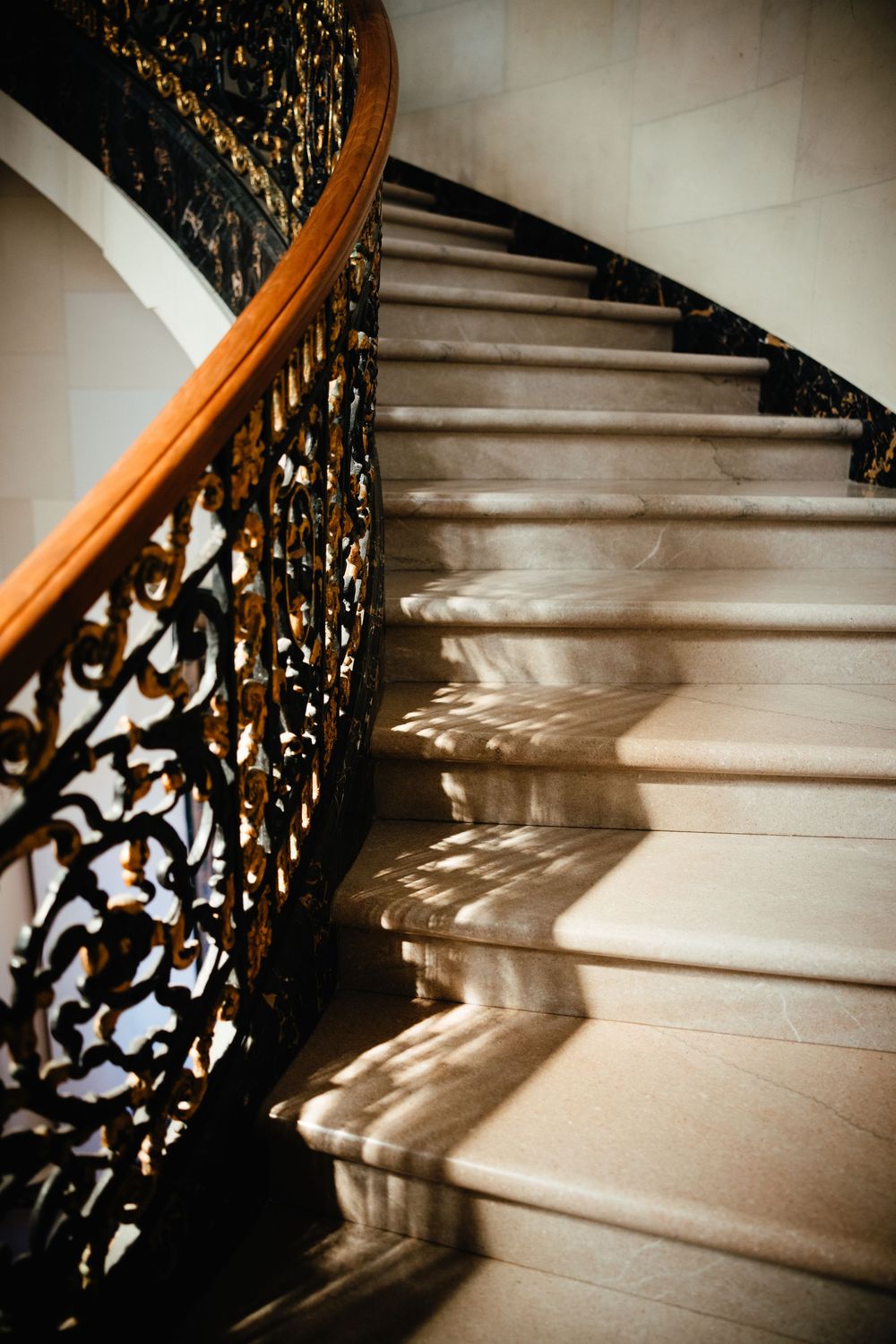 Elegant curved marble staircase with ornate black and gold wrought iron railing casting dramatic shadows in sunlight.