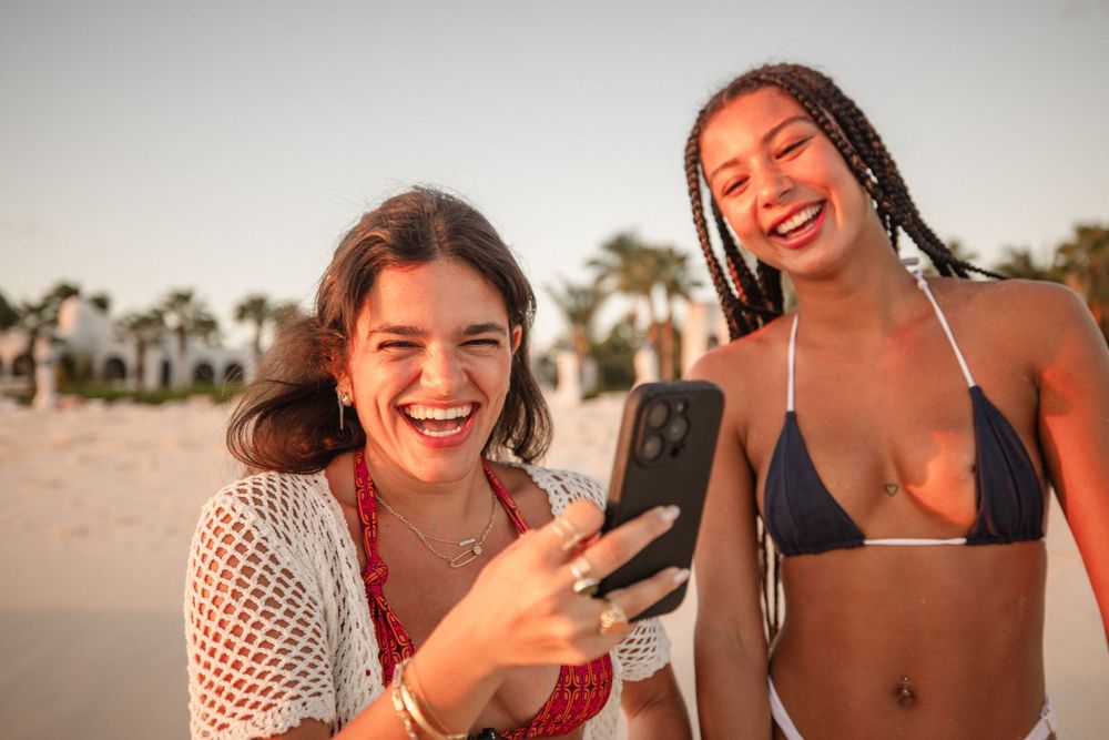 Two young women in bikinis laughing and looking at a smartphone on a sunny beach with palm trees.