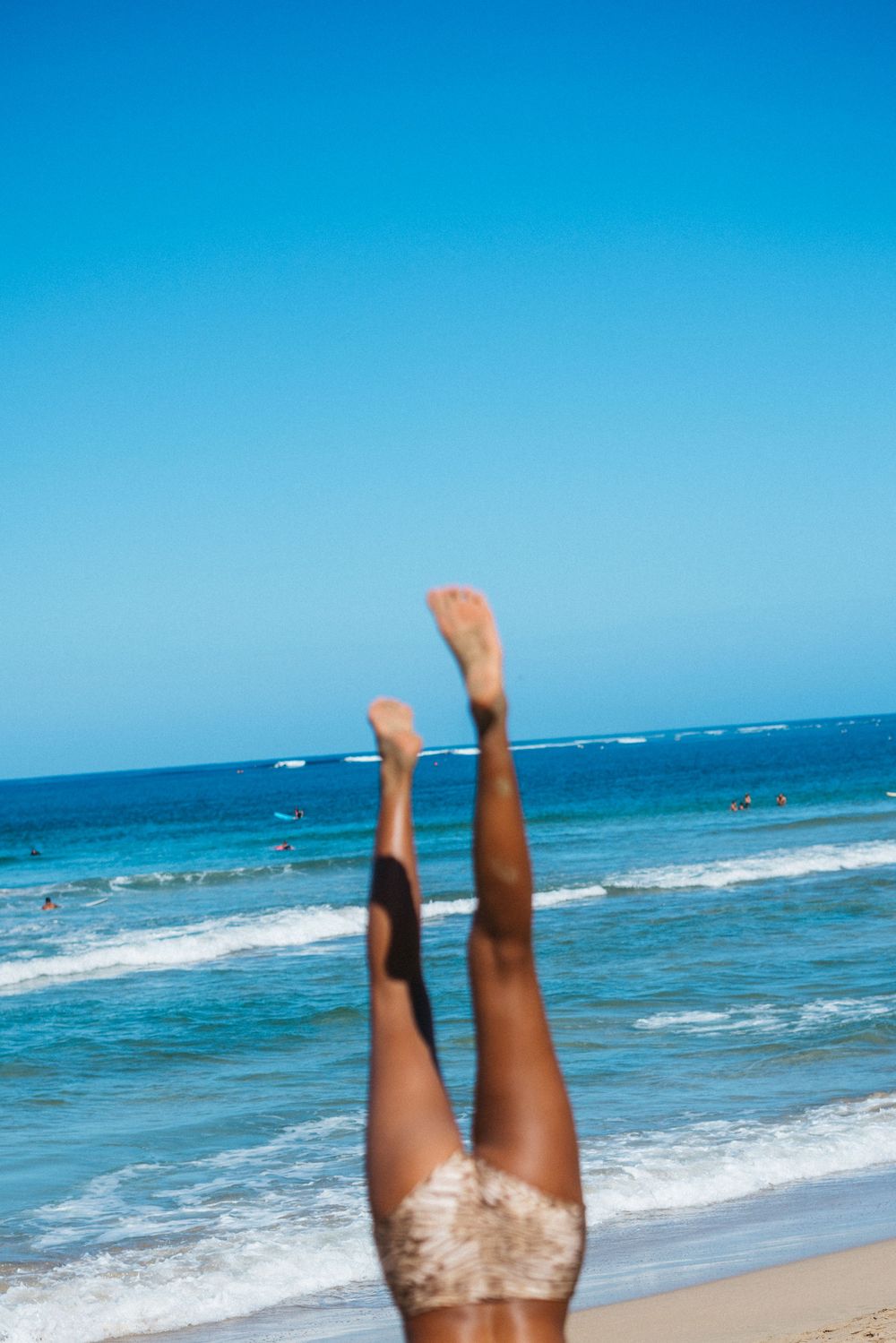 Person doing a handstand on a sunny beach with blue ocean waves and clear sky in the background.