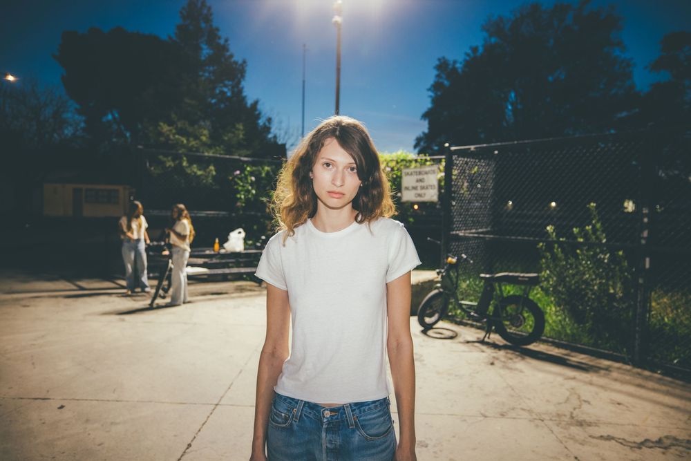 Young woman in white t-shirt and jeans stands on urban street at dusk with trees and people in background.