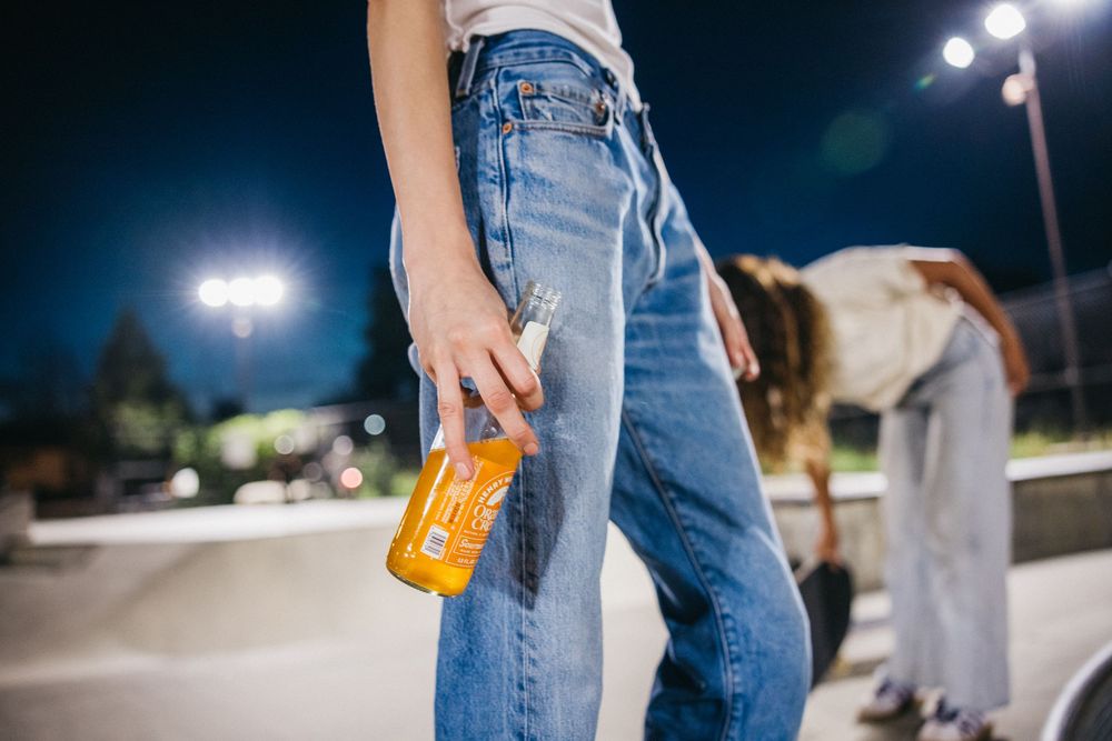 Person in blue jeans holding an orange bottle at a skate park at night with bright lights in background.