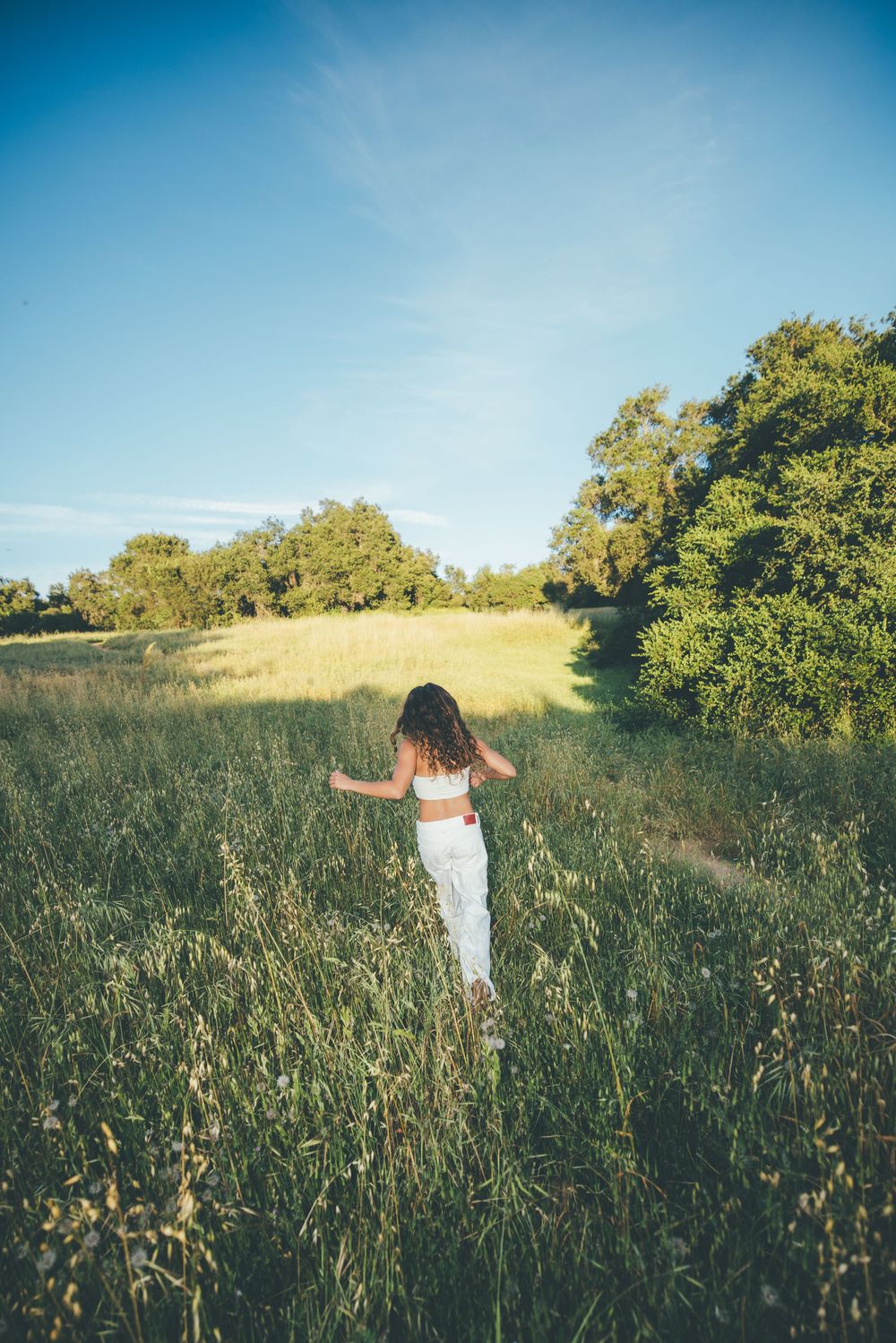 Young woman in white outfit walking through tall green grass field with trees and blue sky backdrop.