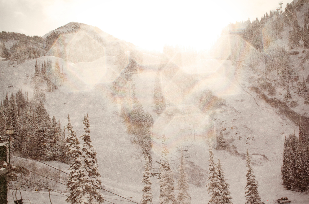 Snow-covered mountain ski resort with pine trees and bright sunlight breaking through winter haze.