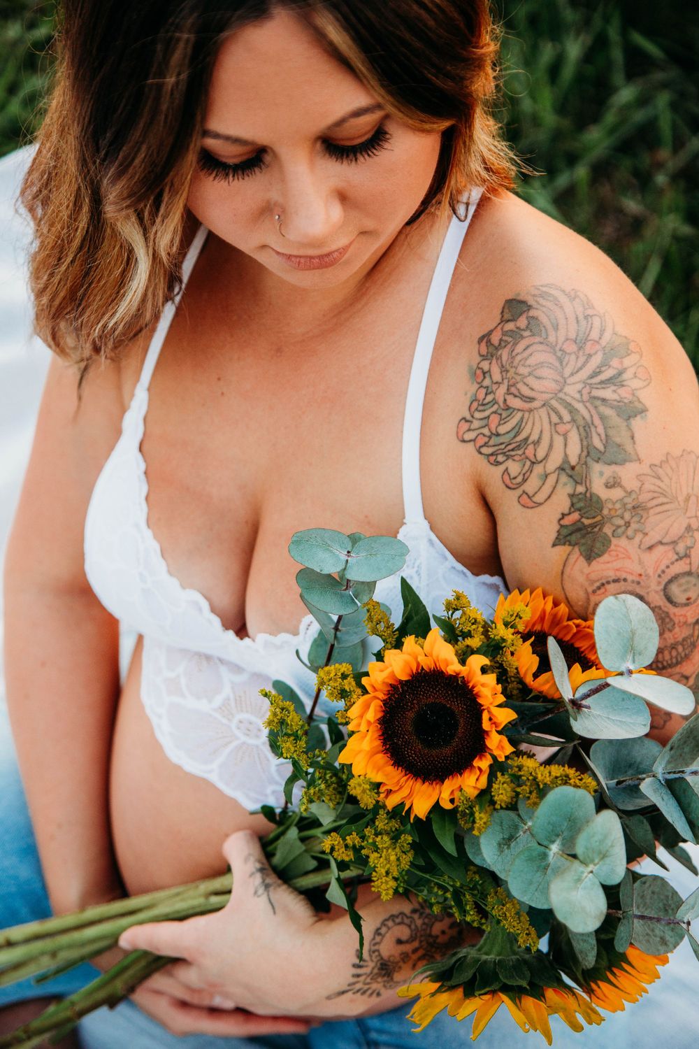 Tattooed pregnant woman in white lace bra holding sunflower and eucalyptus bouquet outdoors.