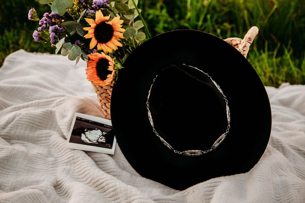 Black wide-brim hat beside ultrasound photo and sunflower basket on white blanket in grass.