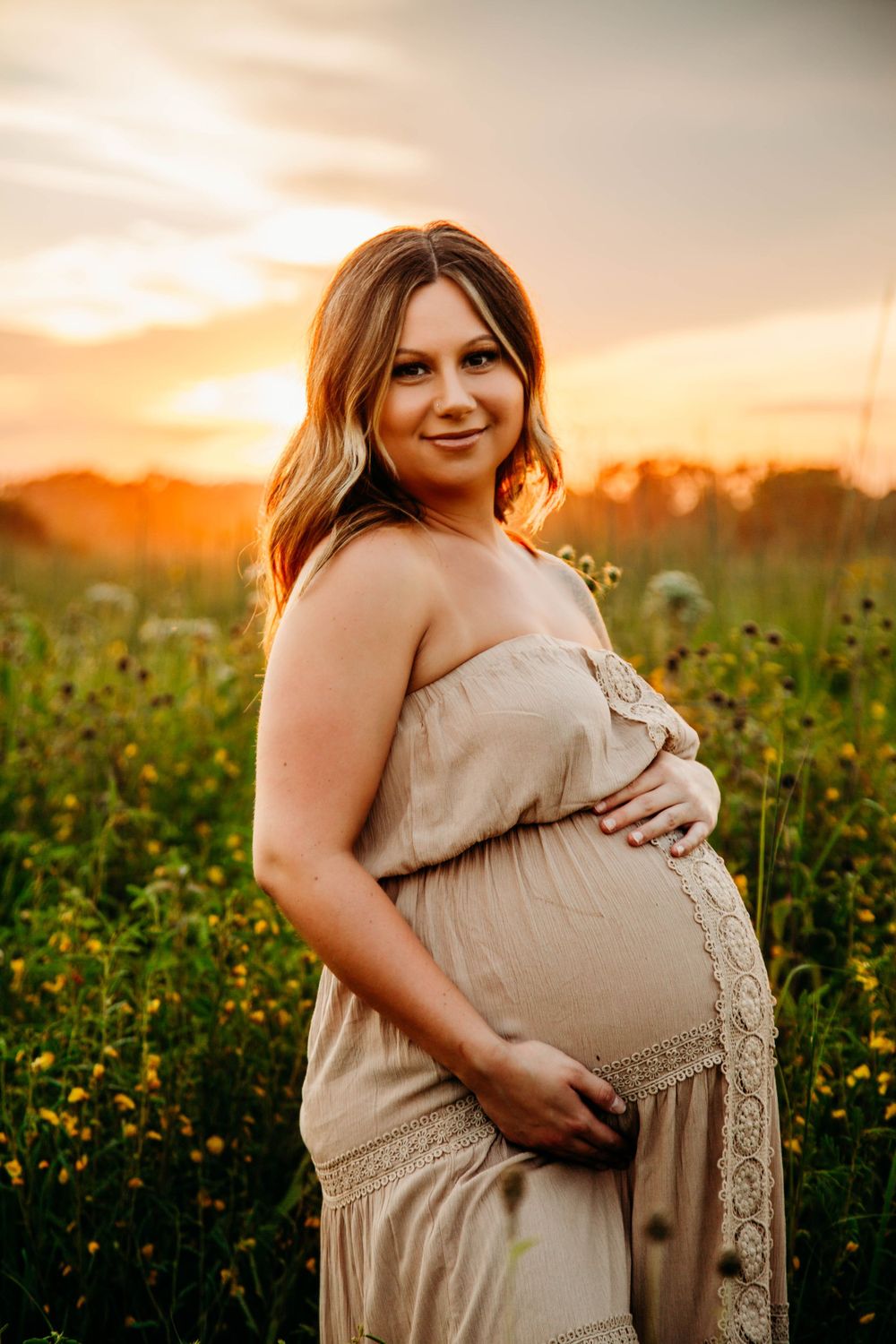 Smiling pregnant woman in beige strapless dress cradling baby bump in golden sunset field.