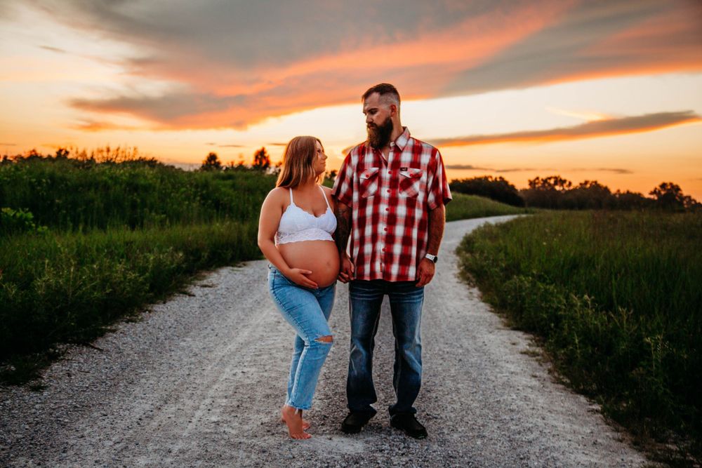 Pregnant couple holding hands on rural dirt road surrounded by green fields at sunset.