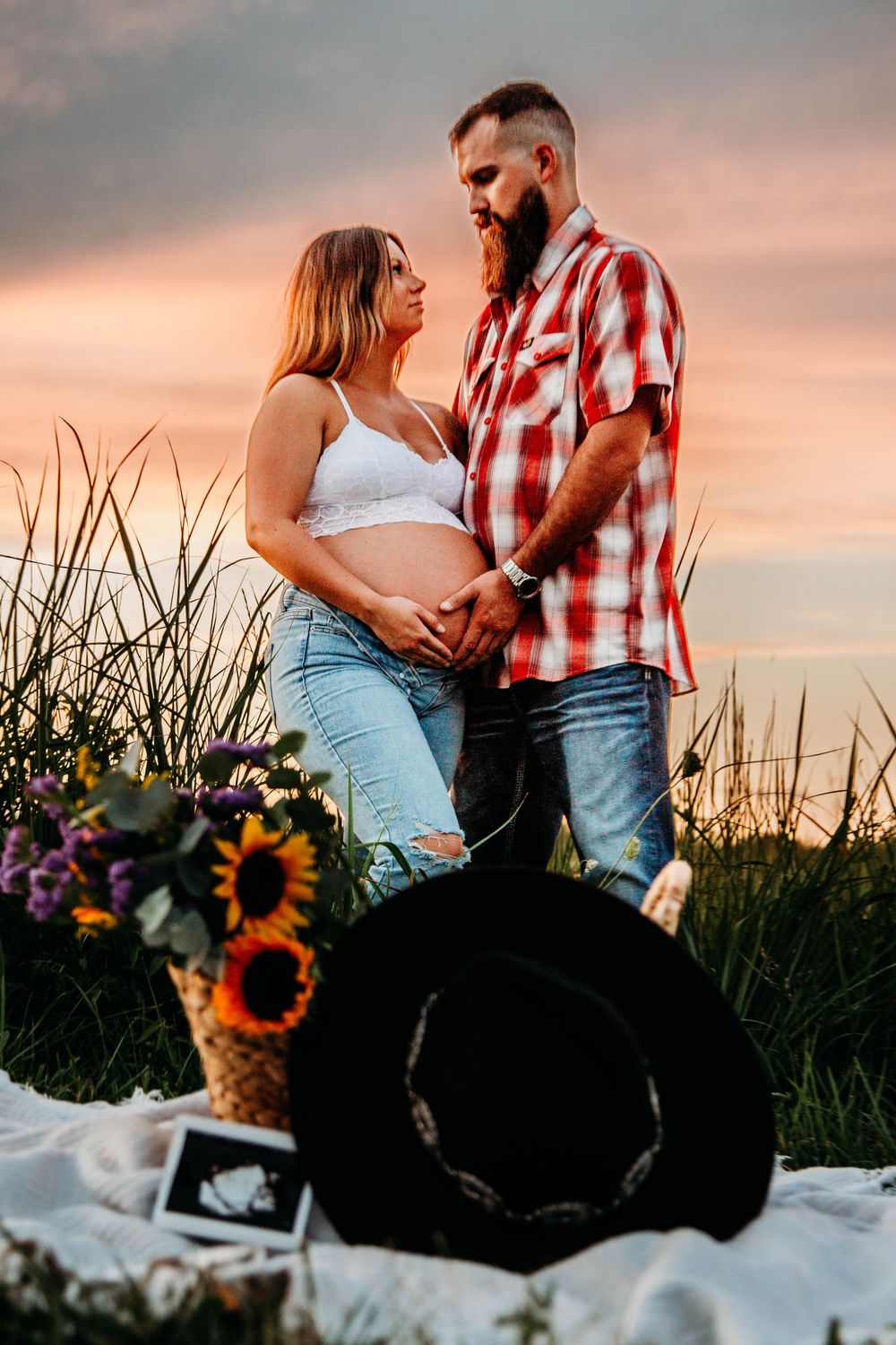 Expecting couple holding hands over baby bump at sunset with sunflowers and ultrasound photo nearby.