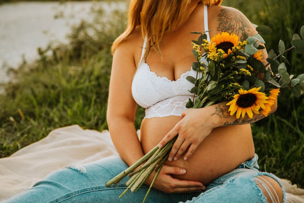Pregnant woman in white lace bralette and jeans holding sunflower bouquet while sitting on blanket outdoors.