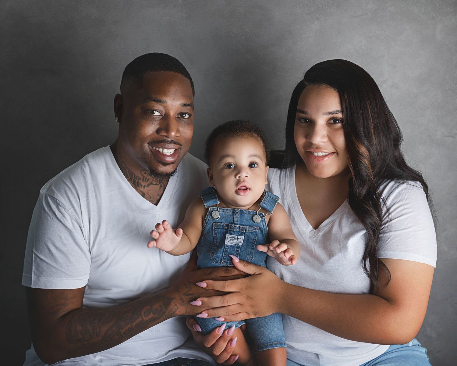 Family wearing white t-shirts poses together for a portrait with their baby in denim overalls.