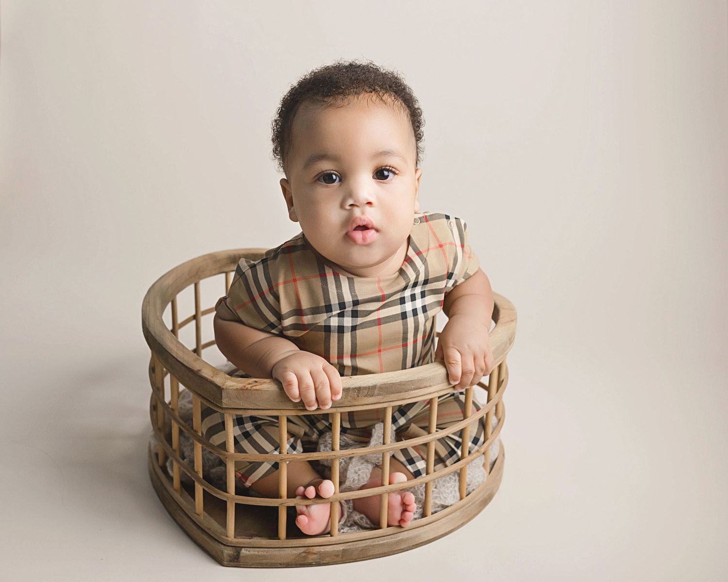 A small child wearing a beige plaid shirt sits in a round rattan basket against a neutral backdrop.