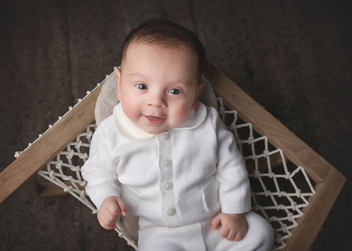 A baby wearing a white sweater sits in a brown wooden chair with a geometric pattern macrame backrest.