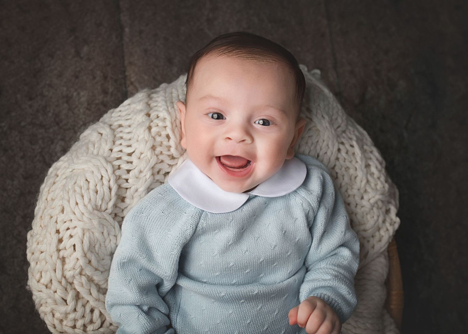 Happy baby wearing a light blue sweater smiles while laying in a cream knit blanket nest during a professional photo session.