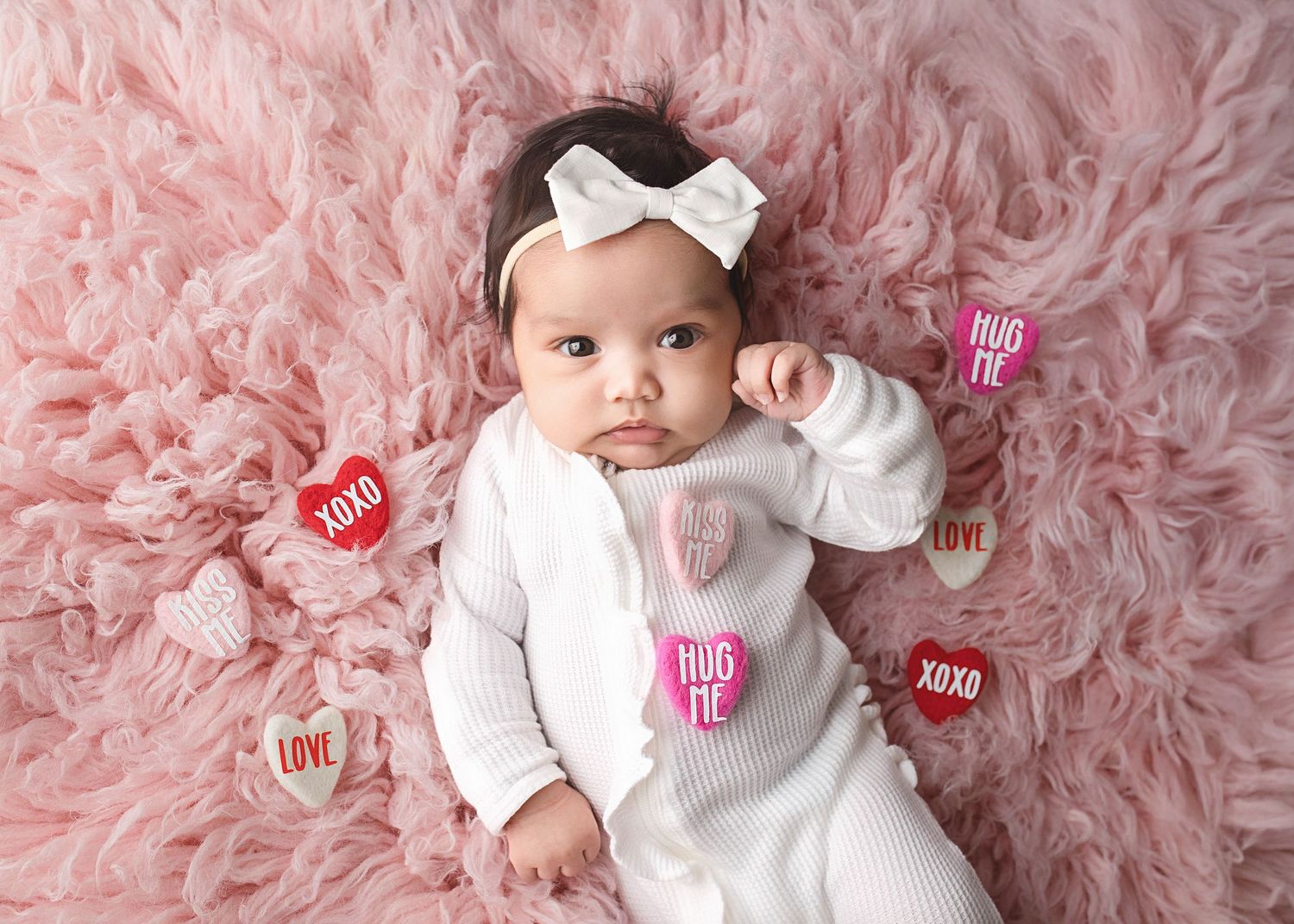 Small baby in white outfit with bow lies on pink plush blanket surrounded by heart-shaped Valentine decorations.