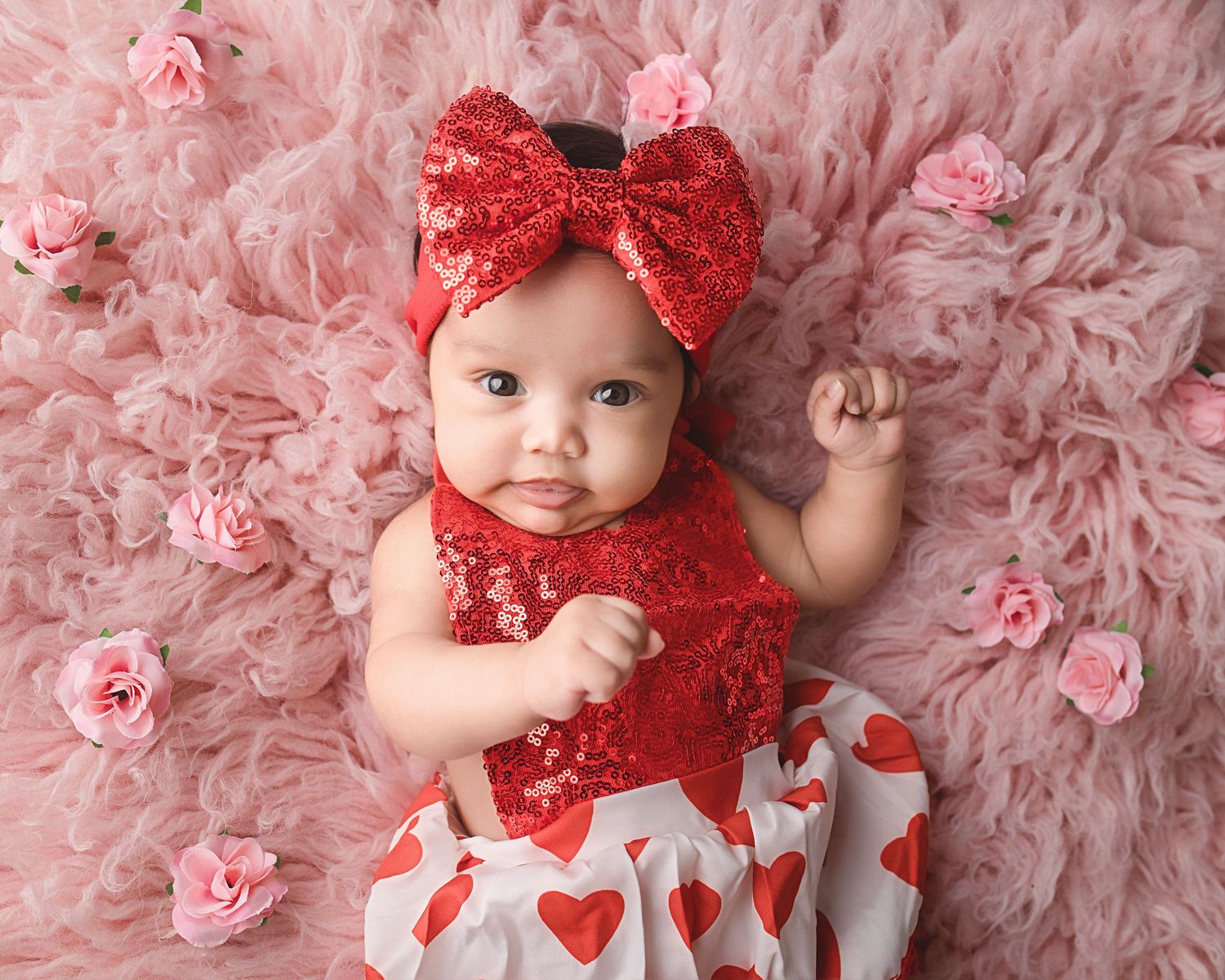 A baby wearing a red sparkly outfit with heart pattern skirt and matching bow laying on pink fabric with roses.