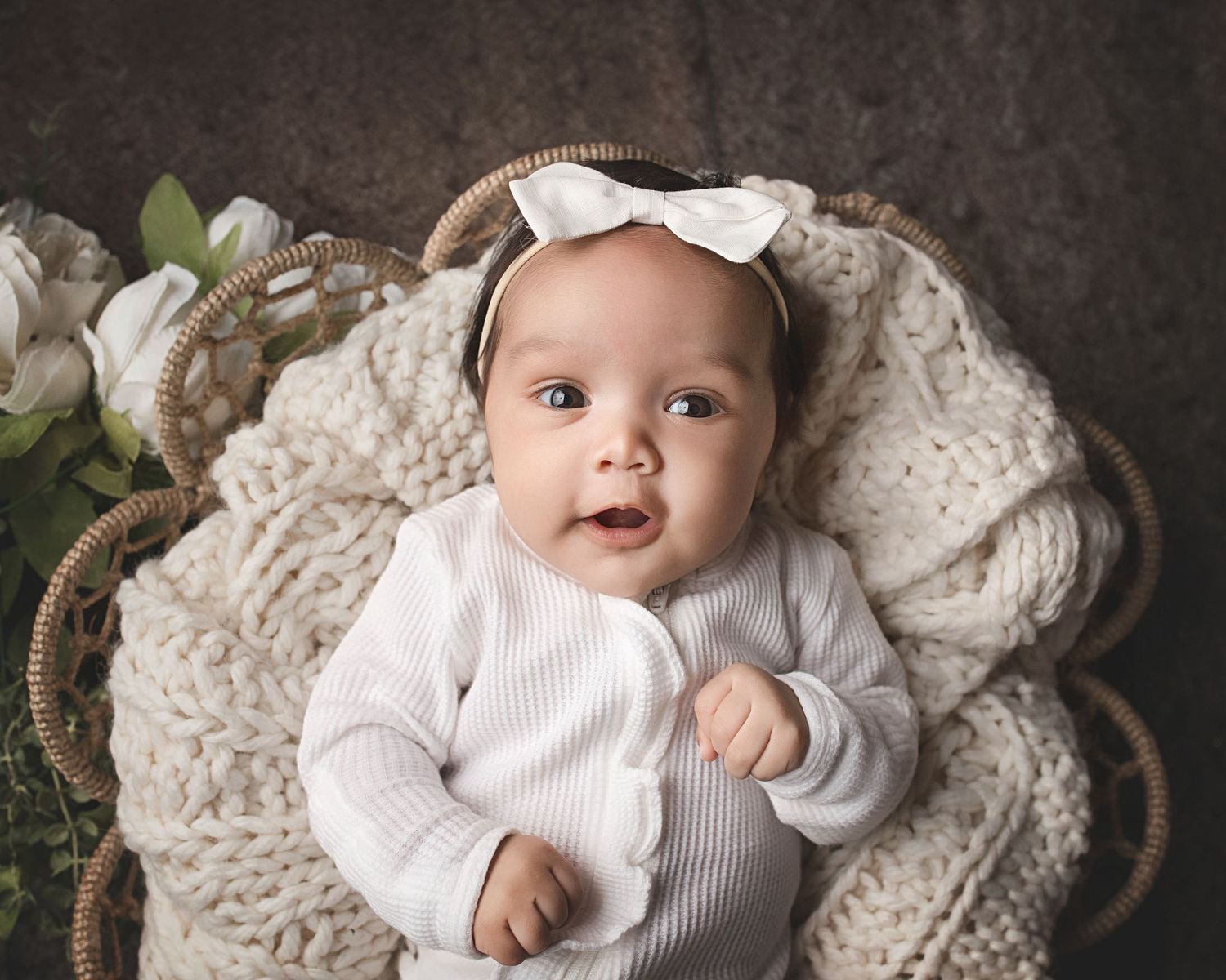 Sweet baby wearing white knitwear and bow lies on cozy knitted blanket with greenery decor in studio setting.