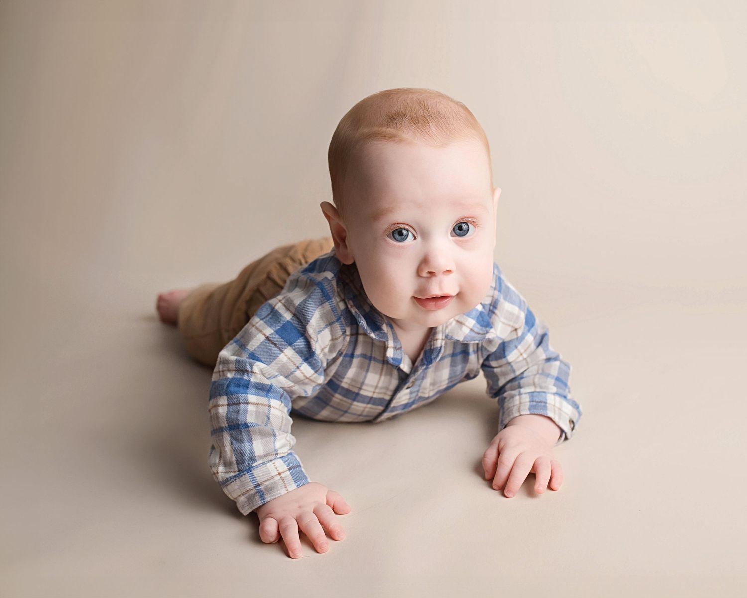 Baby lying on beige backdrop wearing blue and white plaid button-up shirt during professional photo session.