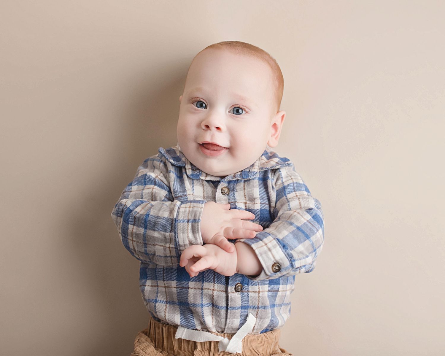 Sweet baby wearing a blue plaid button-down shirt and khaki pants smiles against a neutral beige backdrop.