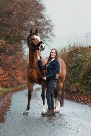 Person in black jacket walking with brown horse on wet autumn path.