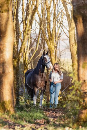 A brown horse and person standing among trees in a wooded area during golden hour.