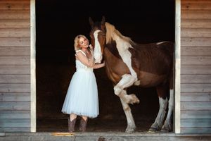 Horse and person in white dress inside a rustic wooden barn with dramatic lighting.