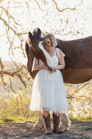 White dress and cowboy boots next to a horse in a dreamy outdoor setting.