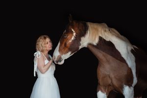 skewbald horse and person in white dress creating artistic silhouettes in a dark barn setting.