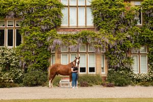 A chestnut horse stands against a vine-covered building facade on a gravel path.