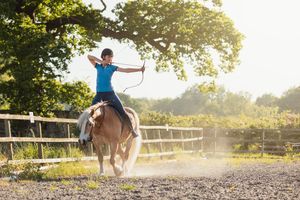 A rider practices horseback riding tricks on a light-colored horse in a dusty arena.