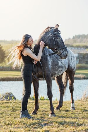 A Appaloosa horse stands by a lake while being gently petted on a sunny afternoon.