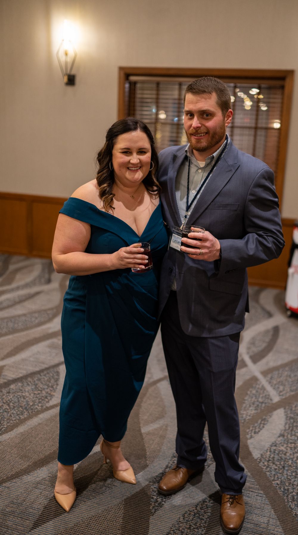 A couple in formal attire poses together at an event, with the woman wearing a teal dress and the man in a navy suit.