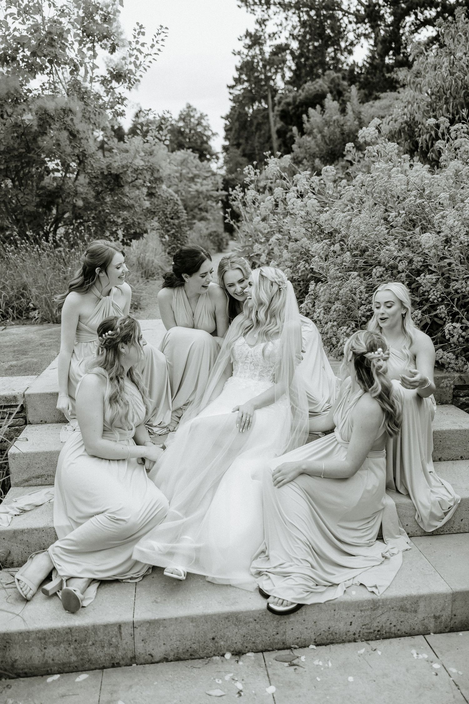 Black and white photo of bridal party in flowing dresses seated on garden steps.
