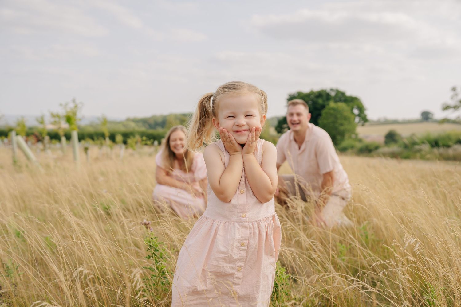 Young girl in pink dress laughing in golden field with parents crouching behind her on a sunny day.