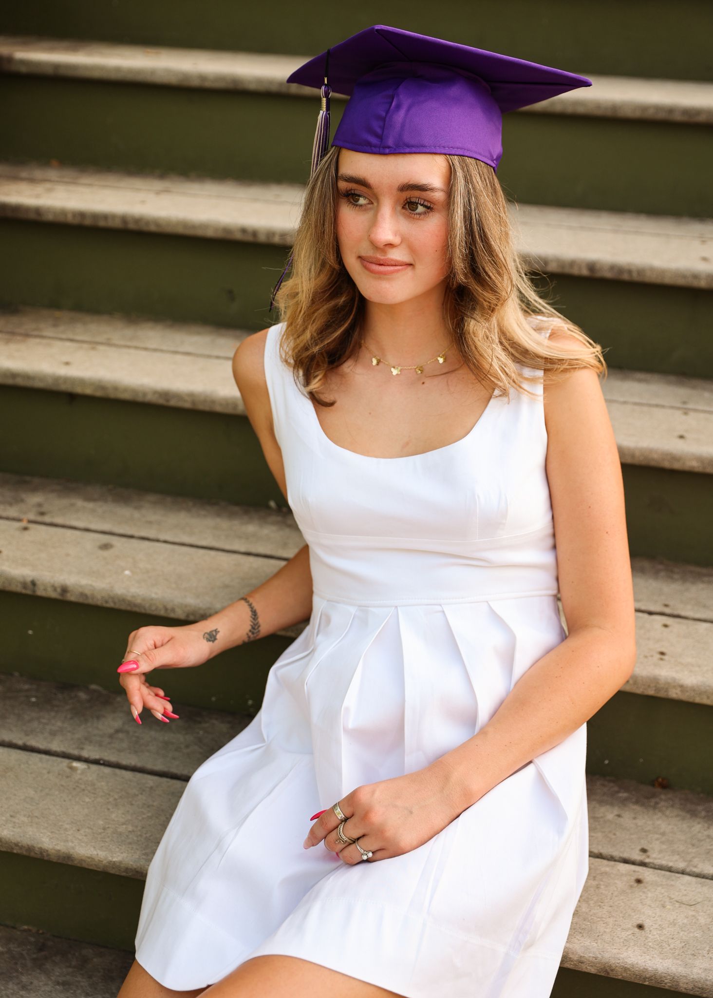 Graduate in white dress and purple cap poses on concrete steps for celebration photos.