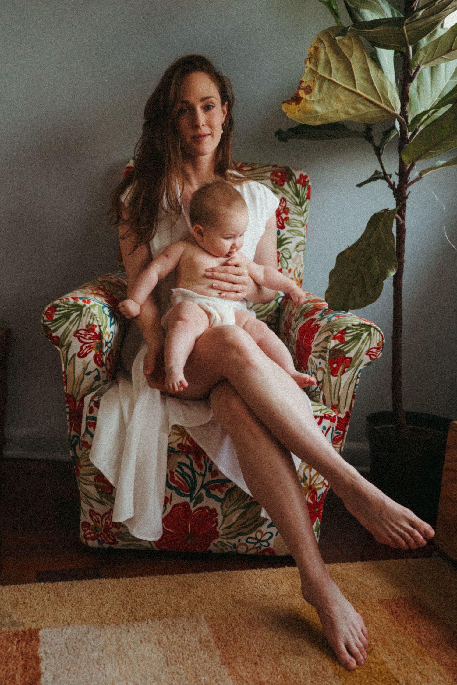 Parent in floral dress holding baby while sitting on floor near houseplant.