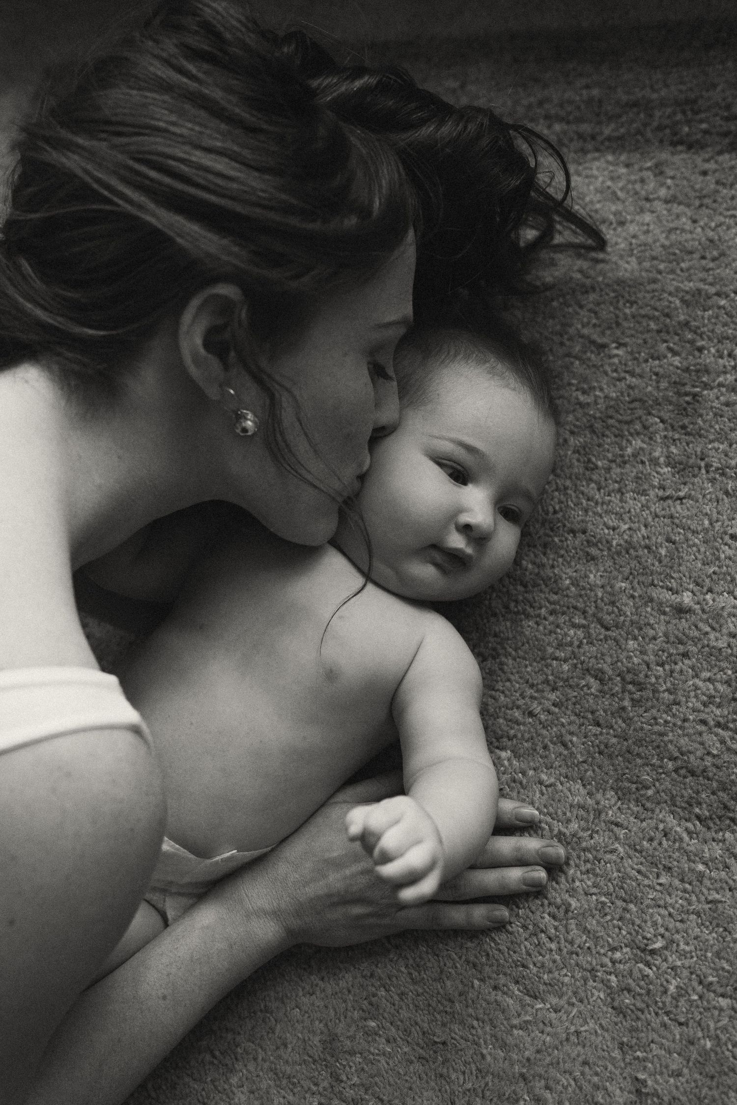 A tender black and white portrait of a mother cuddling with her infant baby on the floor.