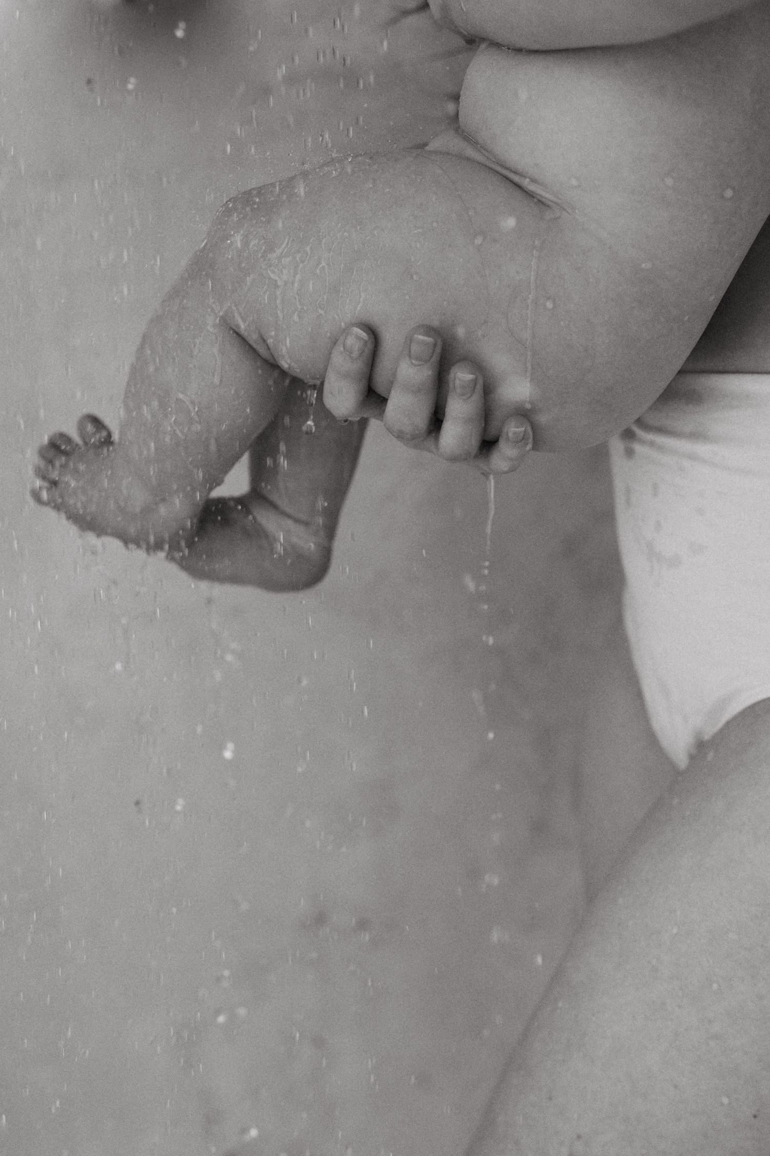 Close-up black and white image of tiny baby feet and adult hands with water droplets.