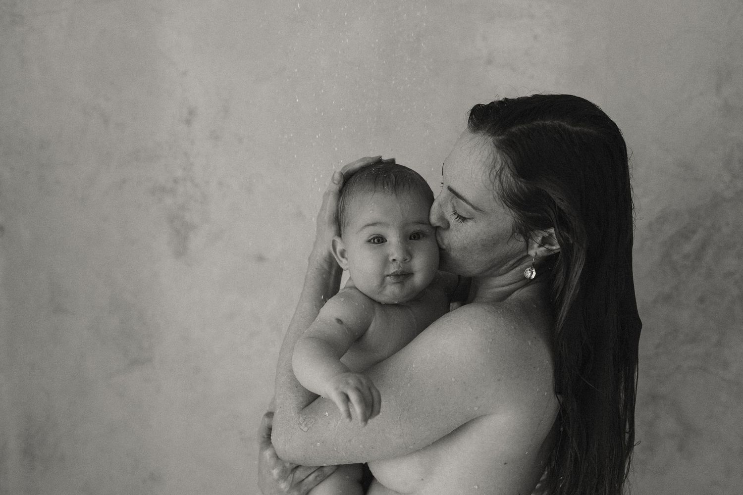 A black and white portrait of a mother kissing her baby against a simple backdrop.