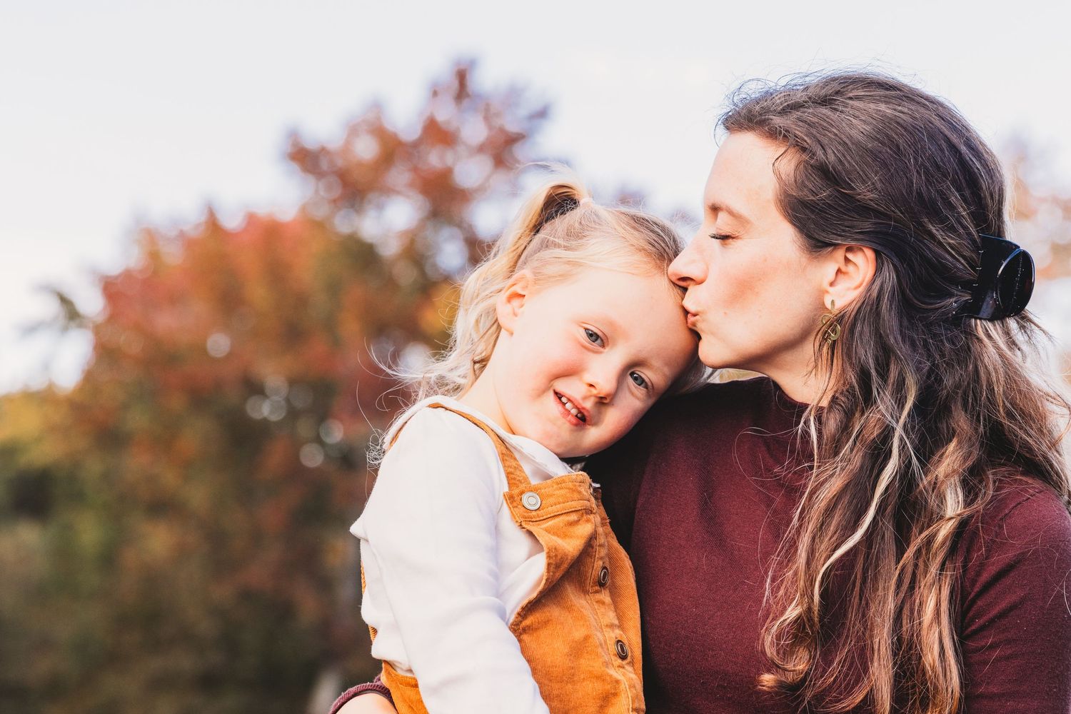 A parent and child share a joyful moment outdoors during autumn with fall foliage in the background at Quiet Waters Park in Annapolis, MD.