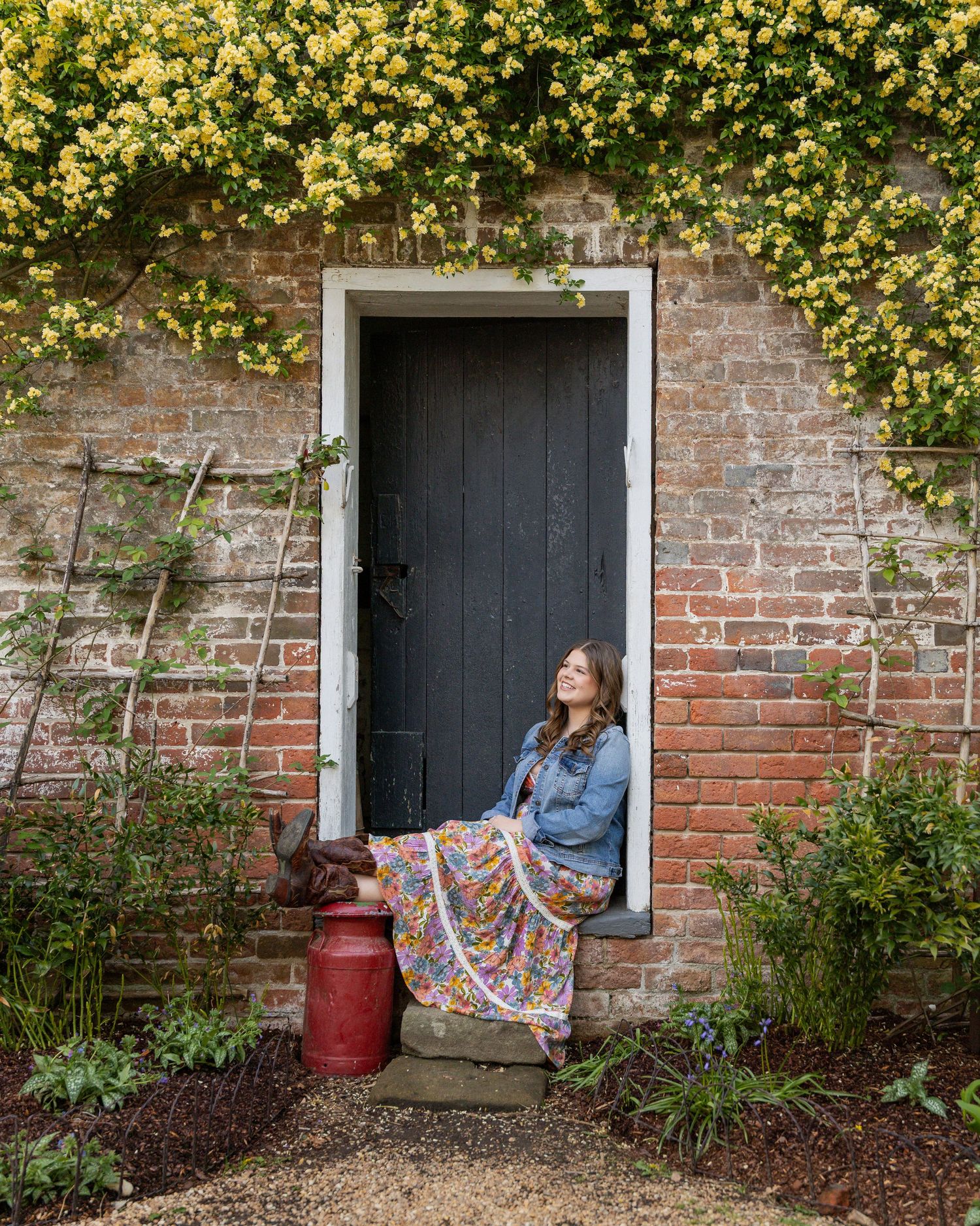 Yellow flowers bloom along a brick wall with black doors and a red barrel in the garden.