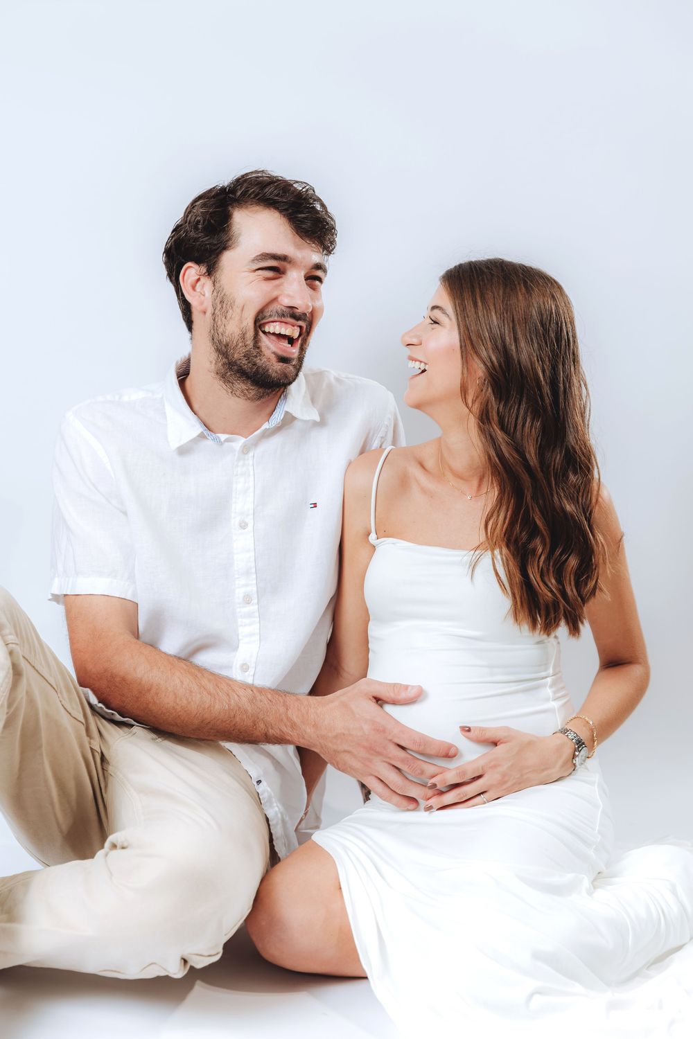 Happy couple sitting together, pregnant woman in white dress cradling baby bump, smiling man in white shirt beside her.