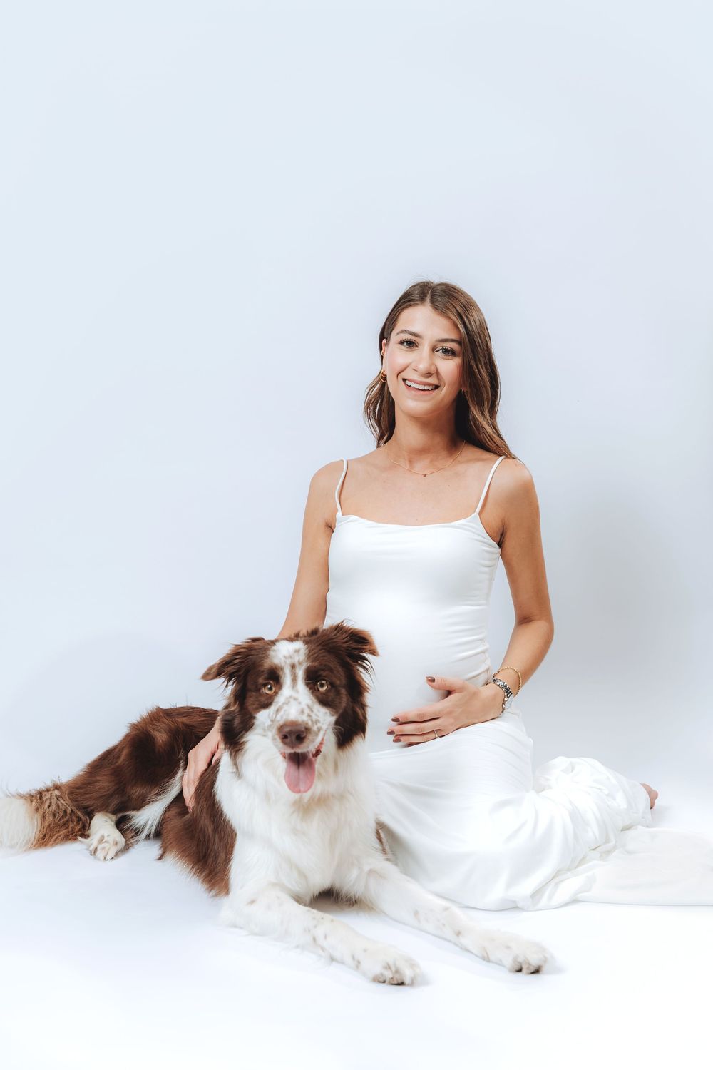 Smiling pregnant woman in white dress sitting with a brown and white Border Collie dog on white background.