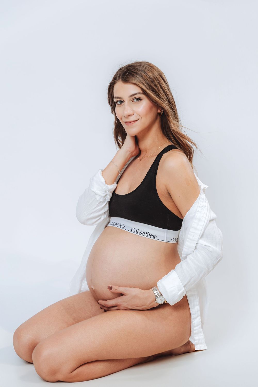 Pregnant woman in Calvin Klein bralette and white shirt sitting on floor cradling baby bump in maternity photoshoot.