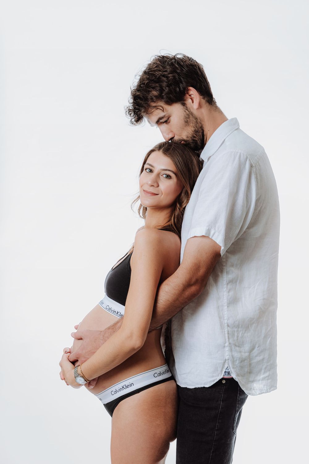 Pregnant couple in Calvin Klein underwear posing together, man embracing woman from behind on white background.
